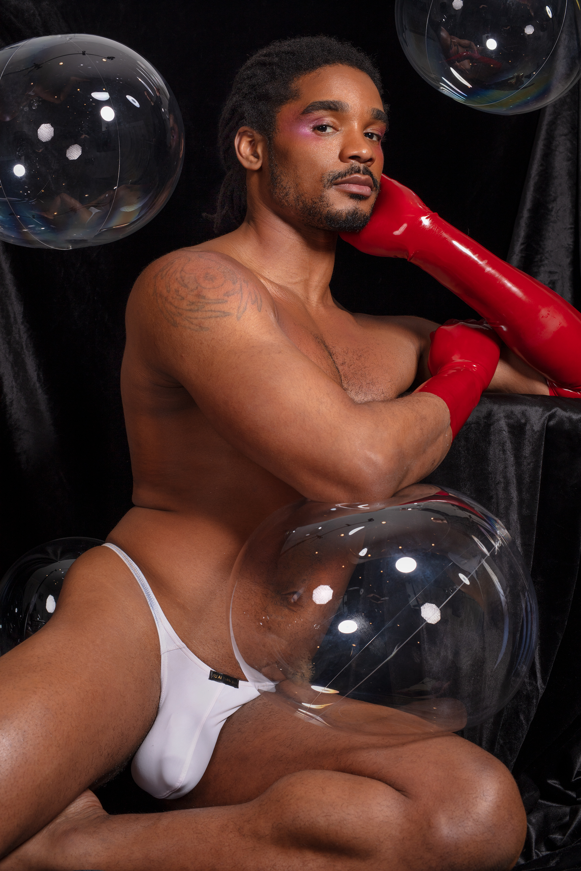 Khalyle suurounded by transparent bubbles, posed leaning with his body outstretched in only a white thong and red latex gloves looking at the camera. 